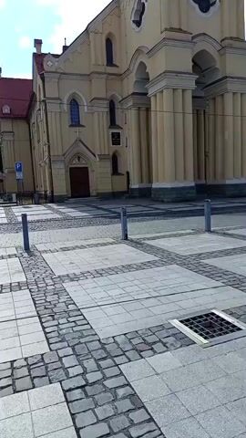 The video shows a time-lapse of a yellow church with a tall steeple and a clock tower under a bright blue sky with fluffy white clouds. The church has a red tiled roof and large arched entrances. The ground in front of the church is paved with large square tiles and cobblestones, with metal bollards and a storm drain visible. The time-lapse captures the movement of the clouds and the changing light, but no people or other significant activity are present. The camera angle is fixed, looking up at the church from a slightly low perspective.