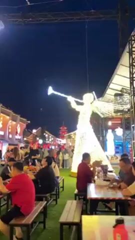 The video shows a crowded outdoor venue at night with tables and seating, suggesting a food market or event. People are seen sitting at tables eating, walking around, and interacting with each other. The area is decorated with bright, festive lights, including a large illuminated angel figure and a building facade outlined in lights. The ground is covered with artificial green turf. The camera moves slightly, providing different views of the scene.