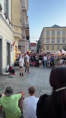 A group of people are gathered in a cobblestone square surrounded by buildings. Two young men, dressed in casual summer attire, are performing a dance or street performance in the center of the square. They are facing a large crowd of spectators who are sitting and standing around them, observing the performance. The buildings surrounding the square are of various architectural styles and colors, with some featuring balconies and awnings. The sky is clear and the lighting suggests it is late afternoon or early evening. The overall atmosphere appears to be lively and festive, with a sense of community engagement.