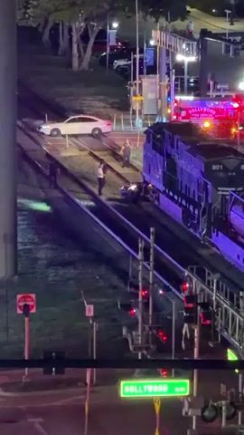 The video shows a train track intersection at night. A train is stopped on the tracks, with its front engine visible. Several people are standing near the train, including what appear to be emergency responders based on a vehicle labeled "HOLLYWOOD FIRE RESCUE". A car is positioned near the train tracks, and it appears to be in the process of being moved or inspected. The area is lit by artificial lights, and there are traffic signals visible. The overall scene suggests an incident involving the train and the car at the railroad crossing.