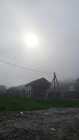 The video opens with a wide shot of a misty, overcast sky with the sun faintly visible through the fog. In the middle ground, a two-story house stands on a hillside, partially obscured by the fog. A power line with a utility pole runs across the frame in front of the house. The foreground is a rough, gravelly area with some green grass on the left.

The perspective then shifts to a close-up view of large, gray rocks scattered on a surface composed of smaller pebbles and some dried vegetation. The camera moves slightly, focusing on the textures and shapes of the rocks and pebbles. The overall impression is of a natural, possibly coastal or riverine environment. No people are visible in the video.