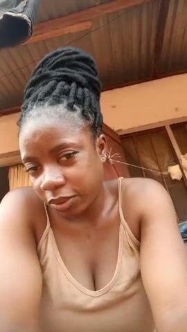 A young woman with dark skin and her hair styled in dreadlocks sits in front of the camera, wearing a light beige tank top. She has a serious expression and sticks out her tongue briefly. The background shows a simple exterior with a corrugated metal roof, cream-colored walls, and glimpses of an open doorway with a person partially visible inside.