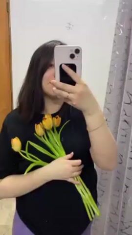 A woman is taking a selfie in a bathroom mirror, partially obscuring her face with a light purple iPhone. She's holding a bouquet of yellow and orange tulips in her left hand. She's wearing a black t-shirt and a light purple garment, possibly pants. The background includes white bathroom tiles, a wooden door frame, and a shower curtain with a gray and white pattern.