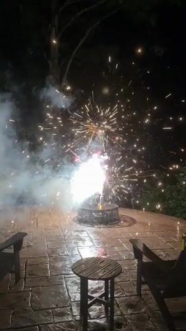 The video captures a nighttime outdoor scene on a paved patio with two chairs and a small table. A fire pit in the center of the patio is actively emitting fireworks. The fireworks produce bright flashes of white and red light, with numerous sparks and smoke billowing outwards. The surrounding trees are visible in the dark background.
