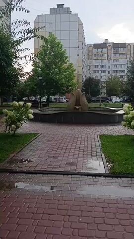 The video captures a view of an outdoor fountain with a tulip-like sculpture in the center. The fountain is surrounded by a stone basin and sits on a paved area with brickwork. Lush green trees and bushes frame the fountain, and in the background, tall apartment buildings are visible, suggesting an urban or suburban setting. The ground is wet, with puddles reflecting the overcast sky, indicating recent rain. The scene is static, with no discernible movement other than the subtle sway of foliage due to a gentle breeze.