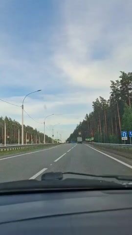 The video shows a first-person perspective of driving down a highway lined with trees. The road is paved with asphalt and has white lane markings. There are pedestrian crossing signs visible on both sides of the road. The sky is blue with scattered clouds. A truck is visible in the distance, driving in the same direction. The interior of a car, including the dashboard and windshield wipers, is visible in the foreground.