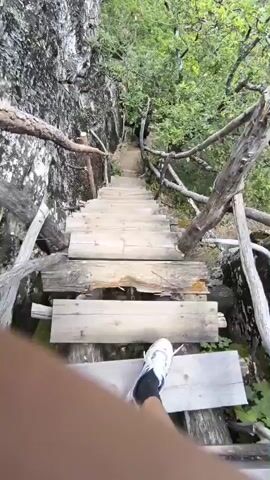 A first-person perspective video shows a wooden staircase descending along a steep, rocky cliff face covered in greenery. The stairs appear rustic and somewhat weathered, with rough-hewn wooden planks and simple wooden railings made from branches. Lush green trees and foliage surround the path, and in the distance, a verdant valley can be seen. The sky above is partly cloudy. A person's legs and feet are visible at the bottom of the frame, indicating they are walking down the stairs.