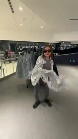 A woman wearing sunglasses, a silver jacket with long fringe, a gray top and gray leggings, and black shoes is standing in a retail clothing store. She is moving her arms and body to make the fringe on her jacket move. There are clothing racks behind her to the left and right. The store has carpeted floors, bright lighting, and black metal railings on a mezzanine level in the background.