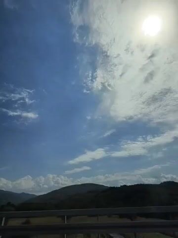 The video shows a bright, sunny day with a blue sky and scattered white clouds. The sun is visible in the upper right corner of the frame, casting a bright glow. Below the sky, there are rolling green hills covered in trees. In the foreground, there is a dark horizontal barrier, possibly a guardrail or fence, running along the bottom of the frame. The overall scene depicts a scenic, natural landscape under a clear sky. There are no people or any discernible actions or interactions taking place. The video appears to be captured from a moving vehicle, possibly a car, given the perspective and the horizontal barrier.