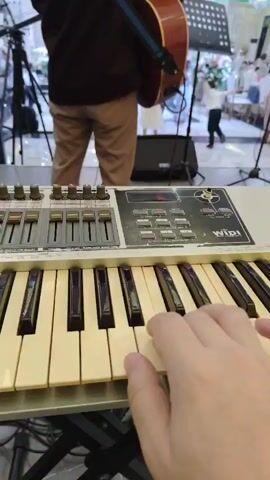 The video is a first-person perspective of someone playing a keyboard, with a person playing an acoustic guitar in the background. The keyboard is white with black and cream keys, and has several sliders and buttons on it. The guitarist, wearing a dark shirt and tan pants, stands on a stage in front of a music stand and speaker. In the background, there is a wedding or formal event taking place with guests dressed in formal attire, including children in suits and dresses. The environment appears to be an indoor venue with decorative lighting. The camera focuses on the keyboard and the player's hands, with the background slightly blurred.