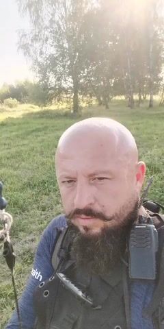 A man with a bald head and a beard is holding a bow in a grassy field. The man is wearing a blue shirt and a vest with a radio attached. The sun is shining through the trees in the background. The man draws the bowstring back to his ear, preparing to shoot an arrow. The arrow is visible on the bowstring. The video ends as the man releases the arrow.