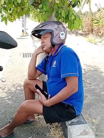 A man wearing a blue polo shirt and black shorts is sitting down with a helmet on his head. He is holding a smartphone in his hand and appears to be resting his head on his hand. He is sitting next to a tree with some vegetation in the background.