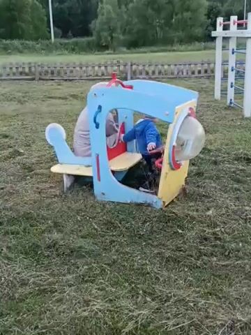 This short video pans across a children's playground in Smolino, Russia. The playground features several pieces of equipment, including a blue and yellow airplane-shaped play structure with a clear bubble cockpit. Two children are visible inside the airplane, one in a blue top and the other partially obscured in a beige top. To the right, a larger play structure with white poles and red and blue bars forms a climbing frame, and a yellow and red pagoda-like structure with a ramp leads up to it. A wooden fence runs across the background, and behind that, a dense line of trees borders the grassy field. The sky is overcast, suggesting it might be late afternoon or early evening. The overall mood is serene and playful, typical of a community playground. No text is visible in the video.