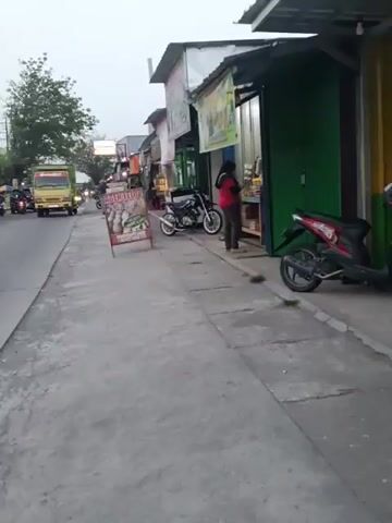 A side-scrolling video shows a sidewalk lined with small businesses. A truck drives by on the left. On the right, a person is standing in front of a shop. Motorbikes are parked on the sidewalk. A sign with pictures of food is placed on the sidewalk in front of a shop. The sky is overcast.