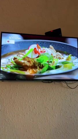 A television screen displays a cooking show from "24Kitchen" featuring a close-up of a bowl of ramen or noodle soup. The soup is garnished with thinly sliced radishes, chili peppers, green onions, and fresh cilantro. A person's hands are visible, carefully adding ingredients to the dish. To the left of the soup bowl, there is a small metal sieve containing pieces of cooked meat, possibly chicken or pork. The background of the cooking set appears to be a wooden countertop with a white placemat under the soup bowl. The television is mounted on a textured beige wall.