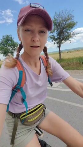 In this media file, a young woman with blonde hair in two braids, wearing a light purple t-shirt, light-colored shorts, a maroon baseball cap, and a fanny pack is riding a black bicycle. She is wearing a backpack with blue and orange straps. The woman is looking at the camera and smiling. The scene is outdoors, with a paved road and a grassy field with some trees in the background. The sky is blue with some white clouds. The time of day appears to be daytime. The weather seems to be warm and sunny. The location appears to be a rural area in Ukraine, specifically the Aromativske rural settlement, judging by the presence of a rural road and fields. The woman's expression suggests a happy and adventurous mood. There is a visible logo on her cap which appears to be "Patagonia".