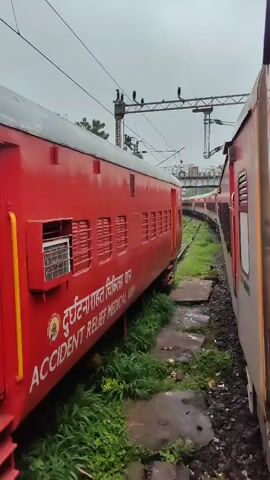 The video is a first-person perspective shot from a moving train. The camera is positioned along the side of a red train car. The red train car has white lettering that reads "RELIEF MEDICAL VAN". To the right of the red train car, another train is visible, which appears to be gray and red. The two trains are running parallel on separate tracks. The tracks are surrounded by green grass and vegetation. In the background, there is a railway bridge and electrical wires overhead. The sky is overcast. The video is taken from a low angle, looking forward along the train tracks. The movement of the train is evident from the blur of the surroundings.