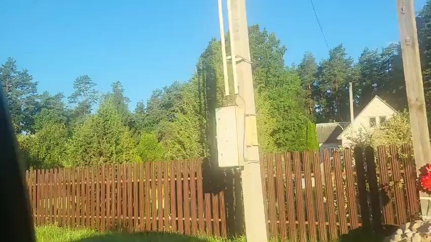 The video shows a residential area with a wooden fence in the foreground and trees in the background. There are two houses visible, one yellow and one brown, with a few smaller structures scattered around them. The houses appear to be in a rural setting, with a lot of greenery and trees surrounding them. The sky is clear and blue, indicating a sunny day. The camera is moving, likely from a vehicle, giving a sense of travel through the area. The overall impression is a peaceful and idyllic rural landscape.