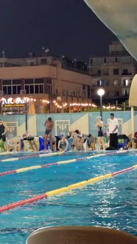 A nighttime shot shows a group of swimmers in a pool, about to start a race. The swimmers are in their lanes, with a man in a white shirt and dark pants standing on the right, appearing to be the official or starter. The pool is illuminated by overhead lights, and the background shows a building with lights and decorations, suggesting it is a public space or a club. The water is disturbed in some lanes, indicating the swimmers are in position and ready to go.