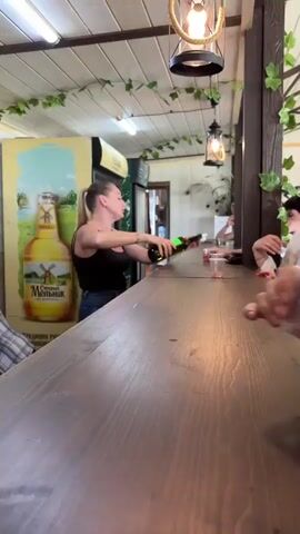 A woman with blonde hair tied in a ponytail, wearing a black tank top and jeans, is seen behind a wooden counter. She is actively pouring a reddish-pink liquid from a dark green bottle into several clear plastic cups held by customers, whose hands are visible reaching across the counter. The woman is speaking and gesturing with her left hand while pouring.

The setting appears to be an indoor bar or a tasting booth. The background features a refrigerator with a yellow and green top section, displaying partial text including "МЕДОВУХА" (MEAD) and "EL...". Above the counter, artificial ivy vines are draped across the ceiling, and two hanging lamps are visible, one designed like an old lantern. A white wall is behind the woman, and a sign with "БАНК" (BANK) is briefly visible. The overall ambiance suggests a casual establishment where beverages are being served.