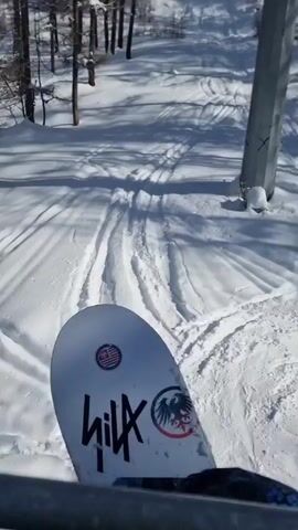 A person rides a ski lift up a snowy mountain, with a Hilx snowboard visible in the foreground. The day is bright, and the sky is blue. Tracks in the snow indicate previous skiers or snowboarders. The mountain is covered in trees. Other skiers or snowboarders can be seen on the lift ahead.