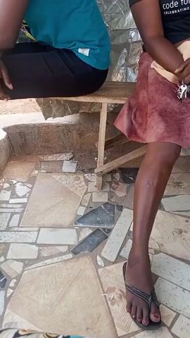 Two people are sitting on a wooden bench. The person on the left is wearing a teal t-shirt and dark pants. The person on the right is wearing a black t-shirt, a reddish-brown skirt, and black sandals. The floor is covered with a mosaic tile pattern. Behind the people is a wall covered in reflective material. The camera appears to be handheld.