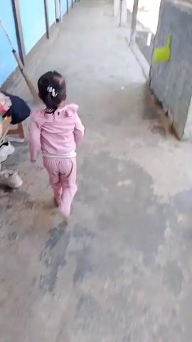 A young child wearing pink pajamas runs down a concrete hallway. The hallway has a covered walkway with pillars on the right side and windows with blue walls on the left. The child's hair is pulled back into a ponytail. There are some people in the distance at the end of the hallway.