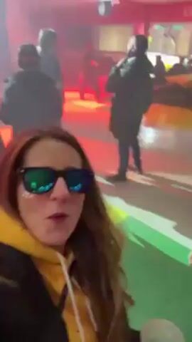 The video shows two people, a woman and a man, taking a selfie in a crowded outdoor area. The woman is wearing sunglasses and a yellow hooded sweatshirt. The man is also wearing a yellow hooded sweatshirt under a jacket. Both are smiling. The background is blurry but suggests a public space with many people and bright red and yellow lights. The camera shakes a lot.