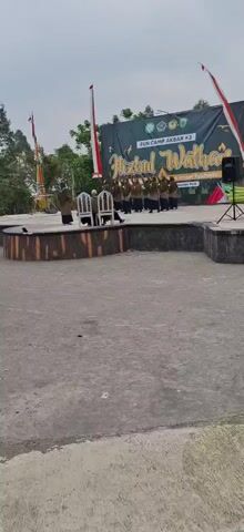 A group of people in brown robes are standing on a stage in front of a banner that reads "FESTIVAL WATHAN FUN CAMP AKBAR #3". Two chairs are on the stage, and one person is sitting in one of them. The stage is outdoors, and there is a crowd of people watching the performance. The weather is overcast.