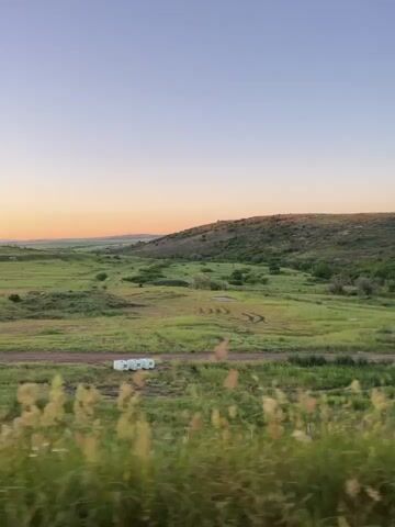 The video shows a daytime, outdoor scene. The foreground is filled with tall grass swaying slightly. Beyond the grass, there is a grassy field with a dirt road running through it. In the distance, a small, white rectangular structure is visible next to the road. The field leads to a gentle hill covered with trees and grass. The sky is a clear gradient, transitioning from a pale blue at the top to a soft orange near the horizon.