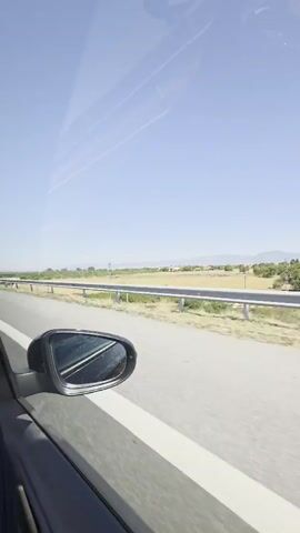 The video shows a view from inside a moving vehicle. The camera is looking out the side window, capturing a bright, sunny day with a clear blue sky. A side mirror of the vehicle is visible in the foreground. Beyond the mirror, a highway or road is seen, with a metal guardrail separating it from a grassy field. In the distance, there are some trees and buildings, and a line of hills under the horizon. There are no people visible in the video, nor any direct interaction between subjects. The overall scene depicts a typical roadside landscape during daytime travel.