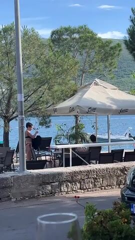 A young woman in a bikini is standing at an outdoor table near a body of water. She is holding a dark garment. There are other tables and chairs around her, some with people sitting at them. Trees and a large umbrella are also visible in the scene. The background shows a body of water with a boat.