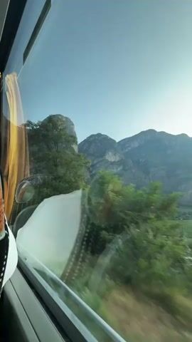 The video is shot from the window of a moving vehicle, likely a bus, looking out at a mountainous landscape. The mountains are rugged and covered in green vegetation. Trees and foliage are visible in the foreground, blurred due to the motion of the vehicle. The sky is clear and blue. Reflections of the vehicle's interior, including a yellow curtain and a white headrest, are visible on the window. In one frame, a triangular road sign with a circular blue sign below it is visible outside. The overall impression is of travel through a scenic, natural environment.