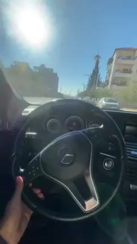 A person is driving a Mercedes Benz through a city street on a sunny day. The shot is from the driver's perspective, focusing on the steering wheel and the road ahead. Buildings are visible on either side of the street, with cars parked along the curb. The sky is clear and bright. The driver's hand is visible on the steering wheel, and a watch is on their wrist.