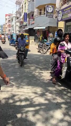 A street scene with several auto-rickshaws and motorbikes. A man in a blue shirt is riding a motorbike in the foreground. In the background, there are buildings with signs and shops. A yellow auto-rickshaw is parked on the right side of the street. Several motorbikes are parked in front of a shop. A woman is riding a scooter with a child behind her. The street is paved and there are shadows cast by the buildings and vehicles. The sky is visible at the top of the frame and appears to be clear.