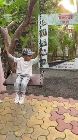 A young boy sits on a swing at an outdoor park. He is wearing a white long sleeve shirt, light wash jeans, and white sneakers. He also has on a baseball cap with a gray pattern. The swing is made of a wooden seat and is suspended by two heavy chains. He holds onto the chains, looking around as he sits on the swing. The playground has an interlocking pavement of red, yellow, and tan. Behind the swing is a sign with an illustration of colorful birds. The backdrop is a lush environment with banana trees and greenery. The lighting is bright, suggesting it is daytime.