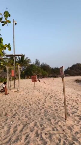 A beach scene unfolds with a clear sky and sand in the foreground. To the left, a metal gate with a yellow fence is visible, accompanied by a security camera mounted on a tall pole. Near the gate, a person sits on the sand. Several orange signs are placed on poles throughout the sandy area. In the background, trees and structures are present, and a few people are scattered across the beach. The overall atmosphere appears calm and the lighting suggests late afternoon.