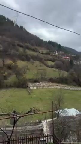 The video presents a scenic view of a small town nestled in a mountainous landscape under a cloudy sky. A green meadow is in the foreground, bordered by a white wall or structure. On the hillside behind the meadow are trees and a few scattered buildings, some of which appear to be residential. A stream or small river flows through the center of the town, with houses lining its banks. Utility lines are visible cutting across the top of the frame. A portion of a wrought-iron railing can be seen at the bottom of the frame, suggesting the view is taken from a slightly elevated position, possibly a balcony or window.