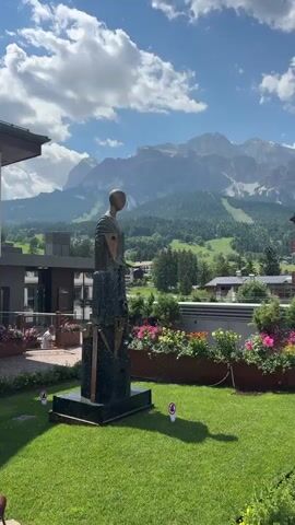 A stationary video captures a view of a garden with a sculpture in the foreground. The sculpture is dark and abstract with geometric elements and a figurative head. Behind the sculpture, there is a low wall with planters filled with colorful flowers, primarily pink and red. In the background, a building with a white facade and a dark roof is visible, along with trees and a valley with houses and ski slopes. Majestic, snow-capped mountains form the backdrop under a cloudy blue sky. The video appears to show sprinklers creating a mist in front of the sculpture.