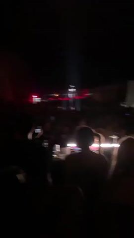 This video appears to capture a live music event or festival at night. A large crowd of people is visible, silhouetted against a stage area. The stage is illuminated by bright spotlights and colored lights, with a prominent beam of light projecting upwards. There are structures that resemble DJ equipment or a sound system on the stage. Some individuals in the crowd are holding up their phones, possibly recording the event. The atmosphere seems energetic, with the lighting suggesting a lively performance. The environment appears to be outdoors, given the darkness of the sky.
