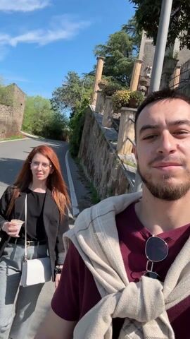A man is holding a phone up to record a selfie video.  A woman is walking on his left. She has red hair and is wearing glasses, a black blazer, gray patterned pants, and a white purse. She is also holding a cup. The man has sunglasses hanging from his shirt and a sweater tied around his neck. They are walking on a sidewalk next to a street. There is a stone wall next to the sidewalk and greenery growing along the wall.  In the background are stairs and a building with columns. The sky is blue.