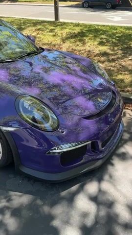 A bright purple Porsche 911 GT3 RS is parked on an asphalt driveway, partially shaded by trees. The car's metallic paint reflects the surrounding environment. The camera is positioned low, focusing on the front passenger side of the vehicle, capturing the wheel, fender, hood, and headlight. In the background, a street with houses and other parked cars is visible. The scene appears to be a sunny day.