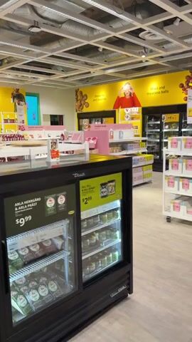 The video pans across an indoor retail environment that appears to be a grocery or food market. In the foreground, a black refrigerated display case is visible, containing various food items such as what appear to be cheeses and prepared meals. Price tags indicate items costing $9.99 and $2.99. To the left of this, a fire extinguisher is mounted on a white pillar. Further into the store, shelves are stocked with drinks, including bottles of wine or sparkling cider, and other packaged goods. A prominent yellow wall in the background features graphics with images of people and text indicating "Sweets and Biscuits" and "Meat and Meatless Balls." The overall environment is brightly lit with fluorescent lighting and has a clean, organized appearance typical of a retail space. No people are directly interacting with the camera or objects in the foreground.