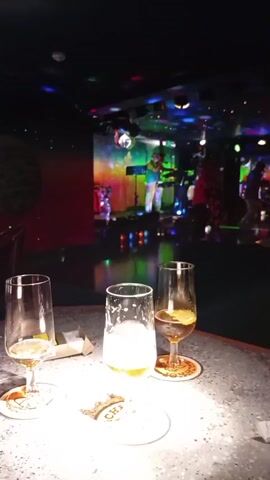 The video shows a dimly lit bar with a stage in the background where a band is performing. In the foreground, there are three glasses of beer on a textured countertop. The band members are visible on stage, with one person playing a keyboard and another holding a microphone. Colorful lights and a disco ball illuminate the stage area. The overall atmosphere appears to be lively and entertaining.