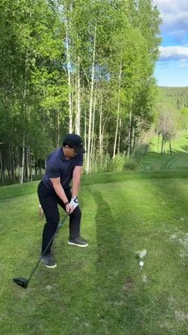 A man wearing a black baseball cap, navy blue collared shirt, and black pants is playing golf on a sunny day. He is swinging a golf club at a golf ball on a tee. The golf course is surrounded by trees. The video cuts to a wide shot of the golf course, showing the green fairway surrounded by trees and a small red building in the distance. The sky is blue with some clouds and the moon is visible.