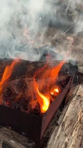 This close-up, vertical video captures a crackling fire in a metal brazier. The subjects are the burning wood, pinecones, and flames, set against a backdrop of dry, possibly sandy or earthy ground, with a larger piece of wood providing a surface for the fire. The scene is a close-up view of a barbecue or campfire setup, likely outdoors given the natural elements and the presence of wood and pinecones.

The activity is the burning of wood and pinecones, indicated by the visible flames, smoke, and glowing embers. The flames are bright orange and yellow, flickering and dancing around the burning materials. Smoke billows upwards and to the left, suggesting a moderate amount of fire and perhaps a slight breeze. Pinecones are visible amongst the burning wood, adding to the rustic and natural feel of the scene. The metal brazier is dark and appears to be well-used, with some rust or discoloration. The surrounding ground is textured and appears dry.

Notable details include the intense heat and vibrant colors of the flames, the wisps of smoke, and the texture of the burning wood and pinecones. There are no visible people, and no text is present in the video. The time of day and weather are not explicitly indicated, but the bright flames suggest active burning, and the presence of smoke and dry ground could imply a variety of conditions. The overall emotion evoked is one of warmth, nature, and perhaps a sense of gathering or outdoor activity. The context provided places this scene in Peski, Russia.