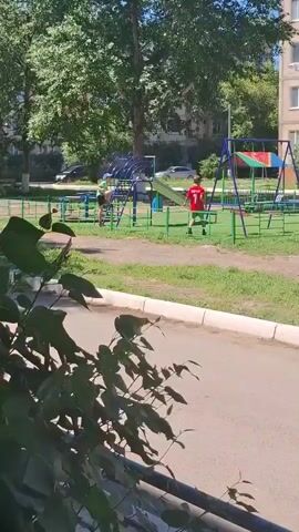 A playground scene on a sunny day. Two boys are visible on a playground. One boy, wearing a green shirt and a blue helmet, is near a blue climbing structure. The second boy, wearing a red shirt with the number 7, is near a green slide. The playground has a green fence and swings. Trees and a building are in the background. The perspective is from behind some foliage, looking towards the playground.