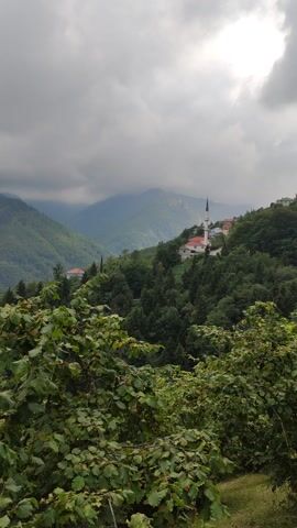 The video shows a landscape with rolling green hills and a cloudy sky. In the foreground, there are lush green bushes. In the background, there are several houses and a building with a tall, pointed structure, possibly a church or a mosque, situated on a hilltop. The camera pans slowly across the scene, revealing more layers of hills fading into the distance. The overall impression is a serene, natural setting with a touch of human habitation.