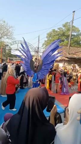 A parade is taking place outdoors on a sunny day. A person in an elaborate costume with large blue wings is walking forward, followed by several other people in colorful costumes. The participants are walking on a red carpet laid out on the ground. A crowd of onlookers, many wearing hijabs, are standing on either side of the carpet, watching and filming the parade with their phones. In the background, there are buildings with tiled roofs and trees. An arched structure with banners is visible in the distance. The overall atmosphere appears festive and celebratory.