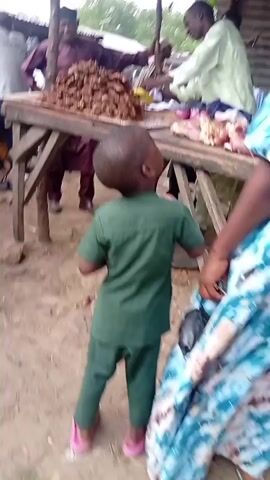 The video depicts a bustling outdoor market scene. A man in a traditional Nigerian outfit, possibly a "babban riga," walks past the camera, his back to the viewer. Another man in a light-colored robe or tunic is seen in the background, interacting with other people or attending to a stall. A young boy in a blue outfit walks past the camera, his attention drawn to something in the distance. The market appears to be crowded, with several individuals moving about. There are various stalls and goods displayed, though the specifics are not clearly discernible due to the camera's movement and focus on the immediate foreground. The ground is dirt, suggesting an unpaved outdoor setting. The atmosphere appears lively and active, typical of a busy marketplace.